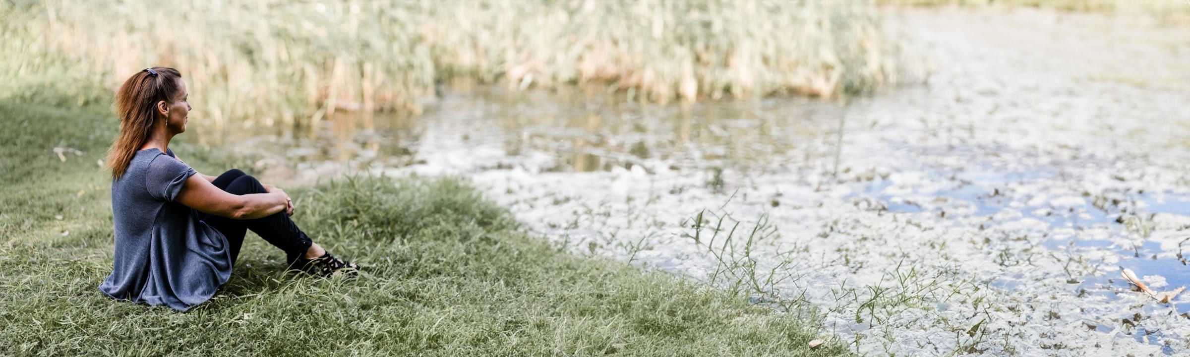Eine Person sitzt ruhig am Ufer eines Gewässers im Gras